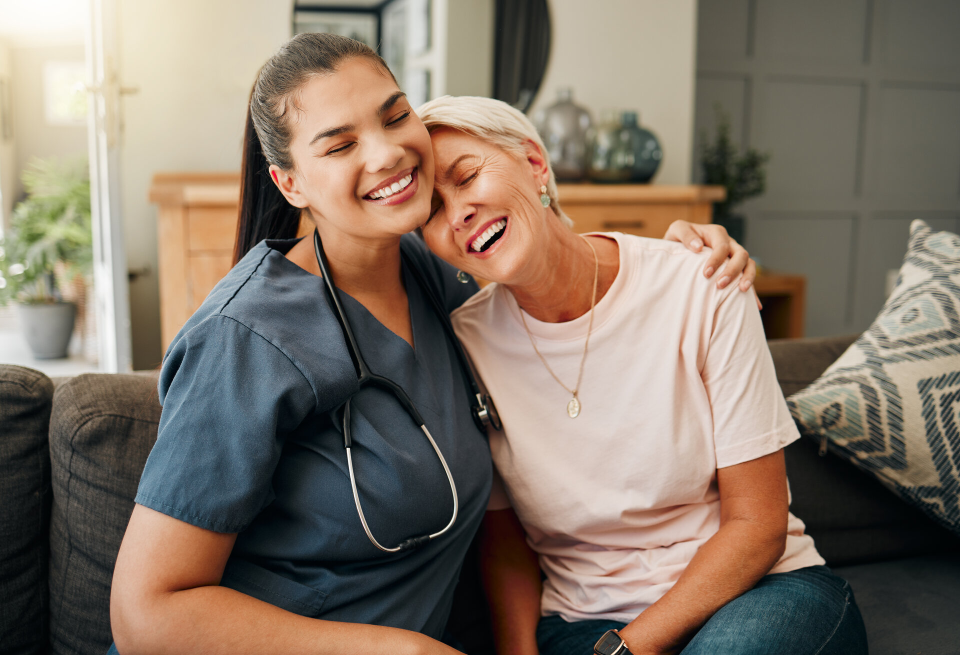 medizinische Betreuerin umarmt lachend eine Seniorin im Wohnzimmer des Hauses